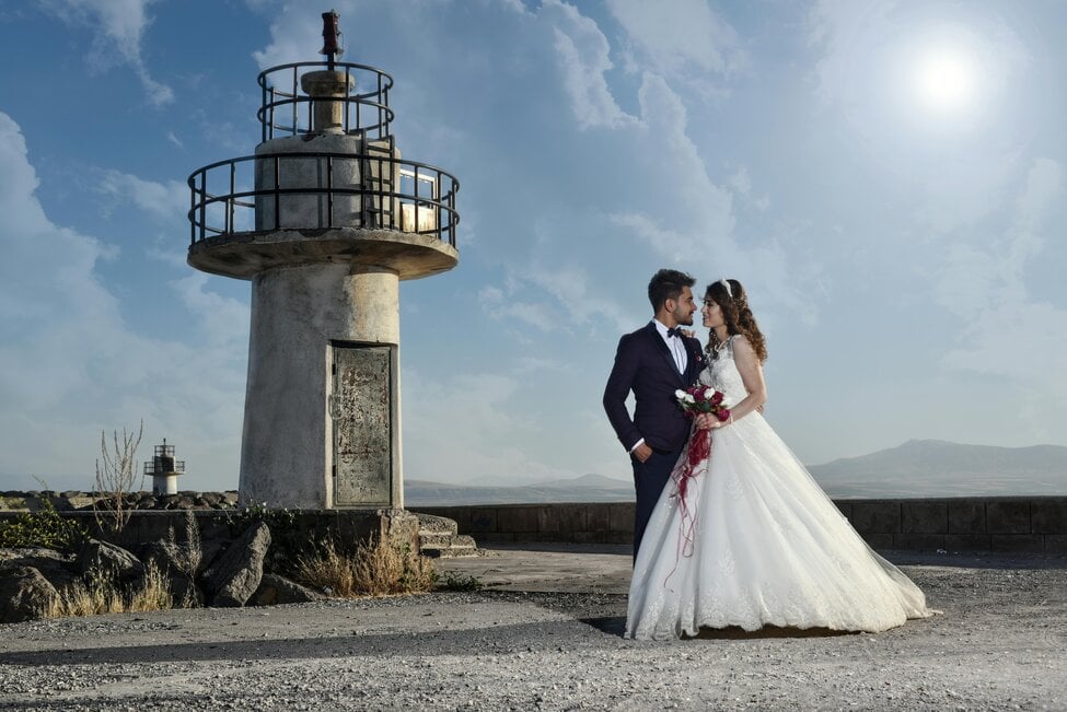 A bride and groom look at each other near a lighthouse on a beach