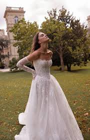 A bride in a flowing off-the-shoulder wedding dress poses gracefully outdoors, surrounded by lush greenery and a historic building in the background.