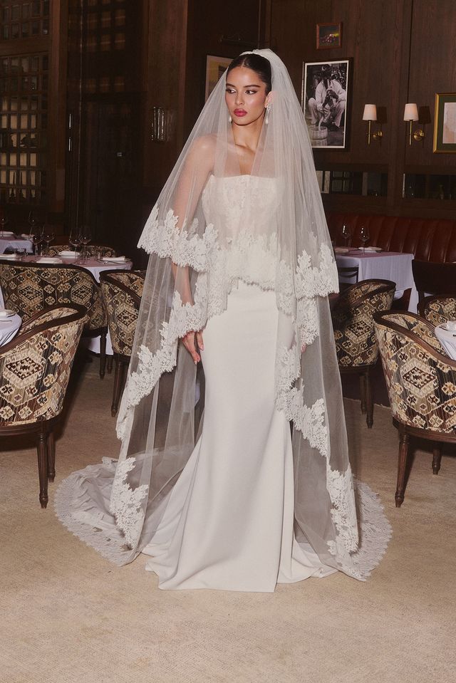 A bride poses elegantly in a sleek, fitted wedding dress paired with a sheer lace-trimmed veil. The upscale restaurant setting enhances the romantic ambiance.