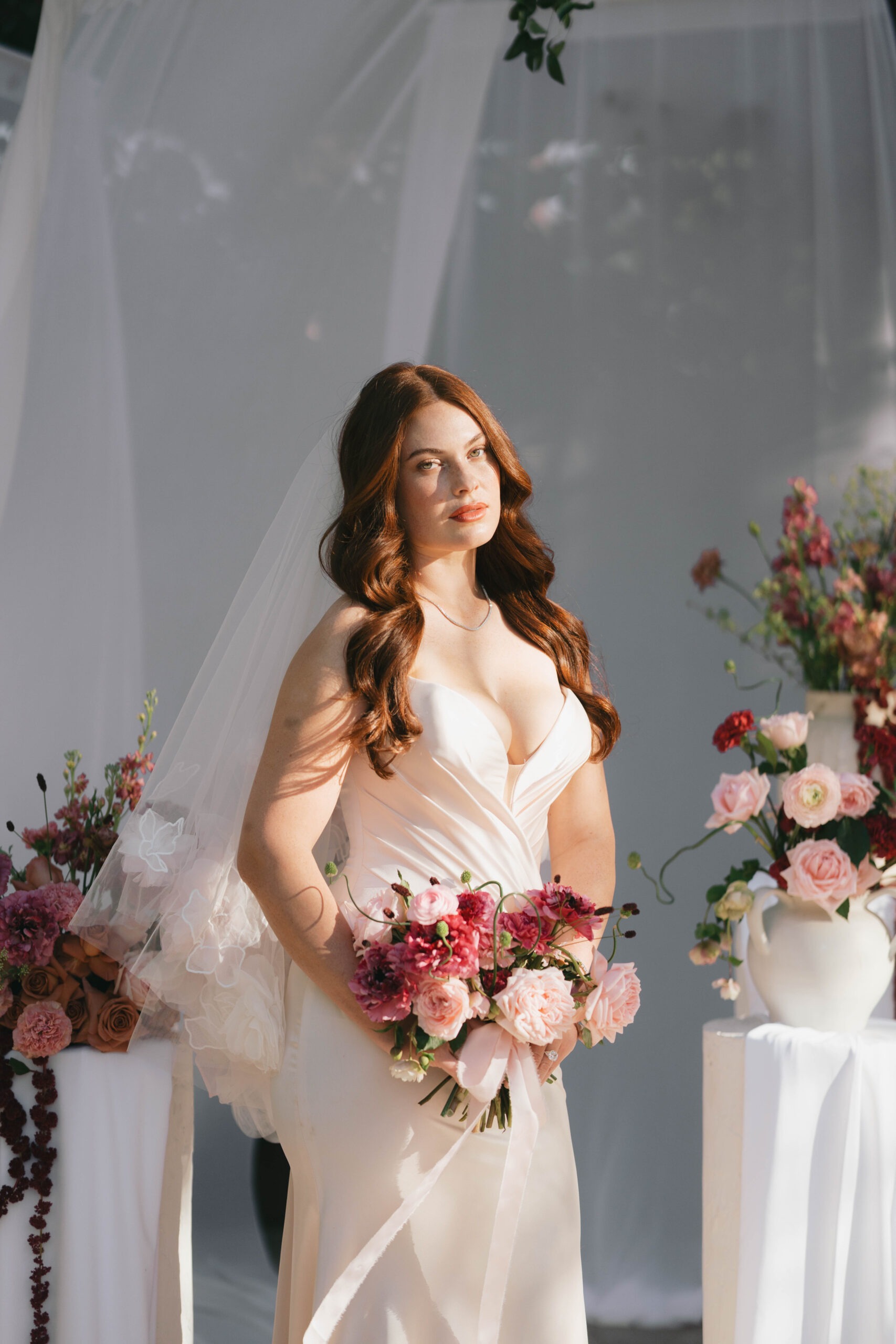 Bride in a flowing gown holds a bouquet of pink and burgundy flowers, standing against a softly lit backdrop with floral accents.