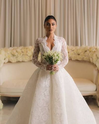 Bride in a lavish white gown with lace details, holding a bouquet of white flowers, stands elegantly in front of floral backdrop, symbolizing elegance and romance.