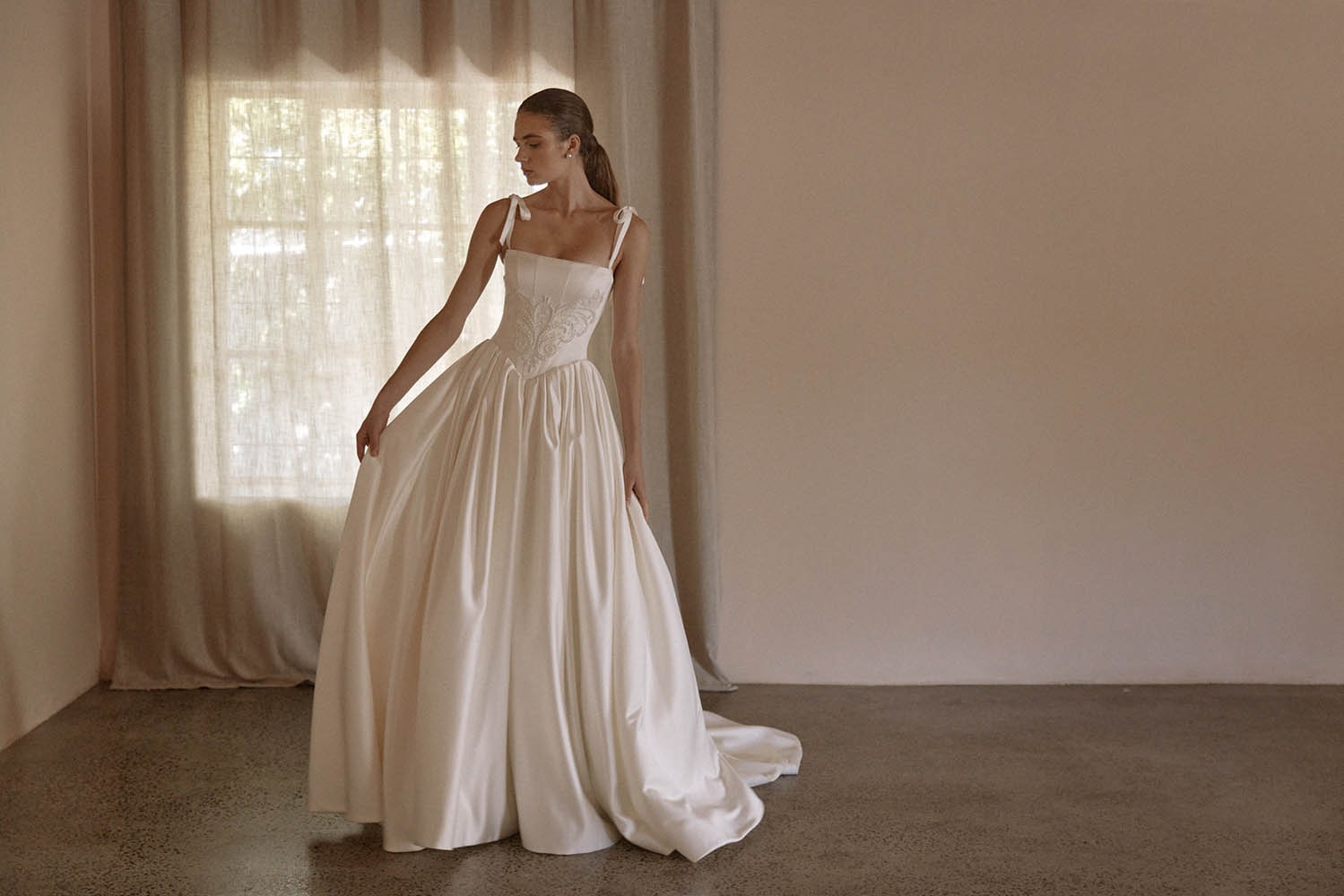 A model showcases an elegant, white wedding dress with a fitted bodice and flowing skirt, set against a softly lit, neutral interior.