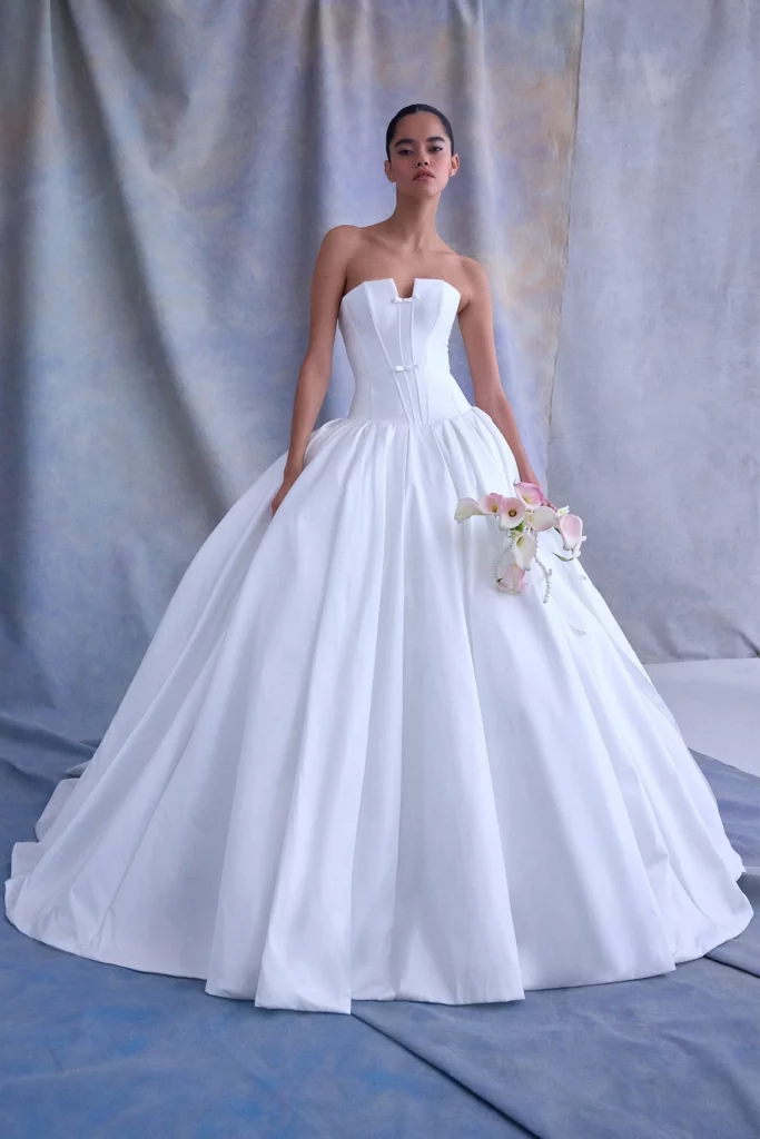 A model showcases a luxurious white gown with a structured bodice and voluminous skirt, holding a bouquet of delicate flowers against a soft backdrop.