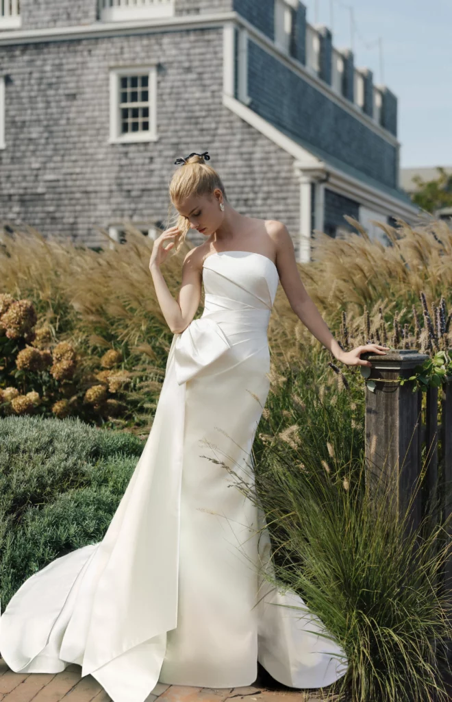 Model in a strapless white gown stands gracefully beside lush greenery and a wooden fence, showcasing elegant bridal fashion in a serene outdoor setting.