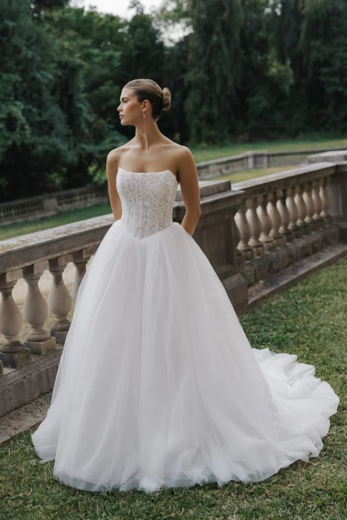 A model poses in a strapless white wedding gown with intricate lace detailing, set against a lush, green outdoor backdrop.