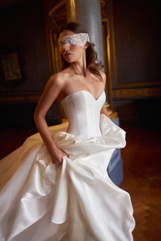 Bridal model in a strapless white gown, holding her flowing skirt, with a lace mask over her eyes, set against an elegant, dimly lit backdrop.
