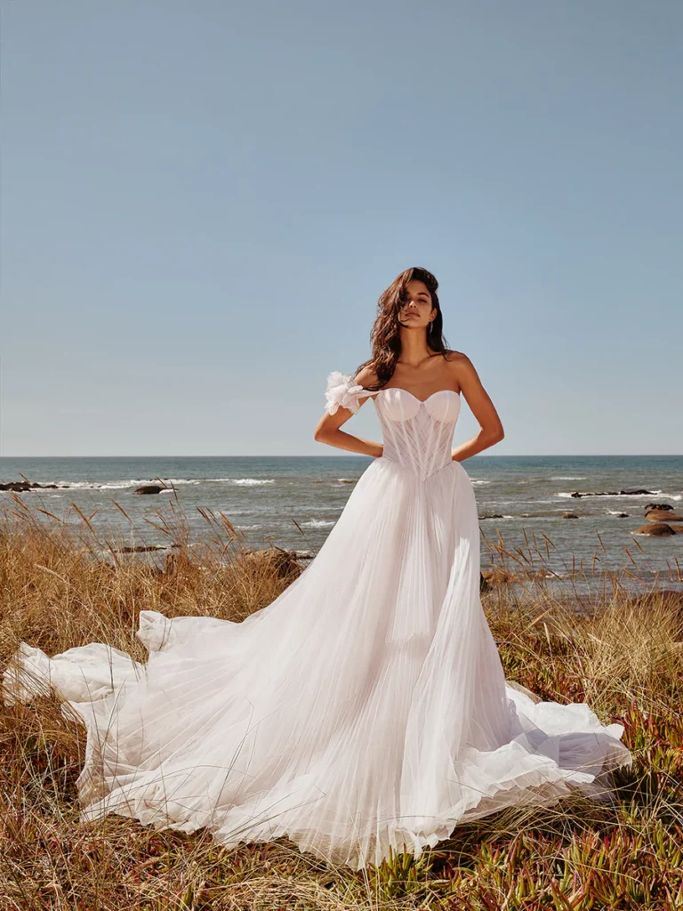 A model poses in a flowing white strapless wedding gown against a coastal backdrop, showcasing the dress's romantic design.