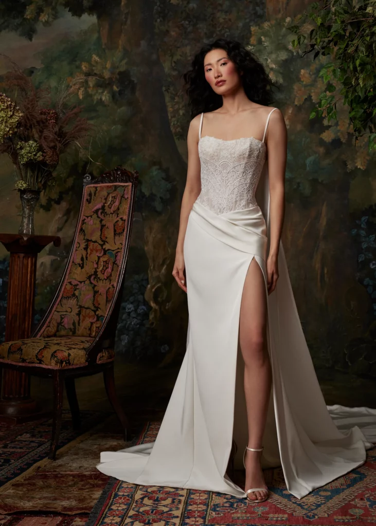 Model in a delicate white gown with a lace bodice and high slit, posed elegantly in front of a painted backdrop and vintage chair.