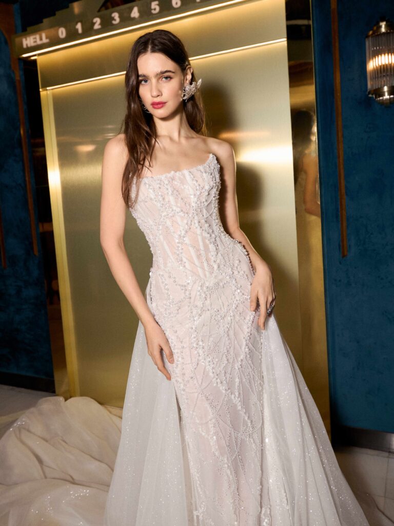 Model in an elegant, beaded white gown with a sheer train poses confidently against a stylish backdrop featuring gold accents.