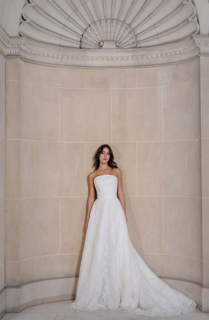 Model poses in a strapless, intricate white lace wedding gown against a cream-colored wall. The elegant setting underscores bridal fashion.