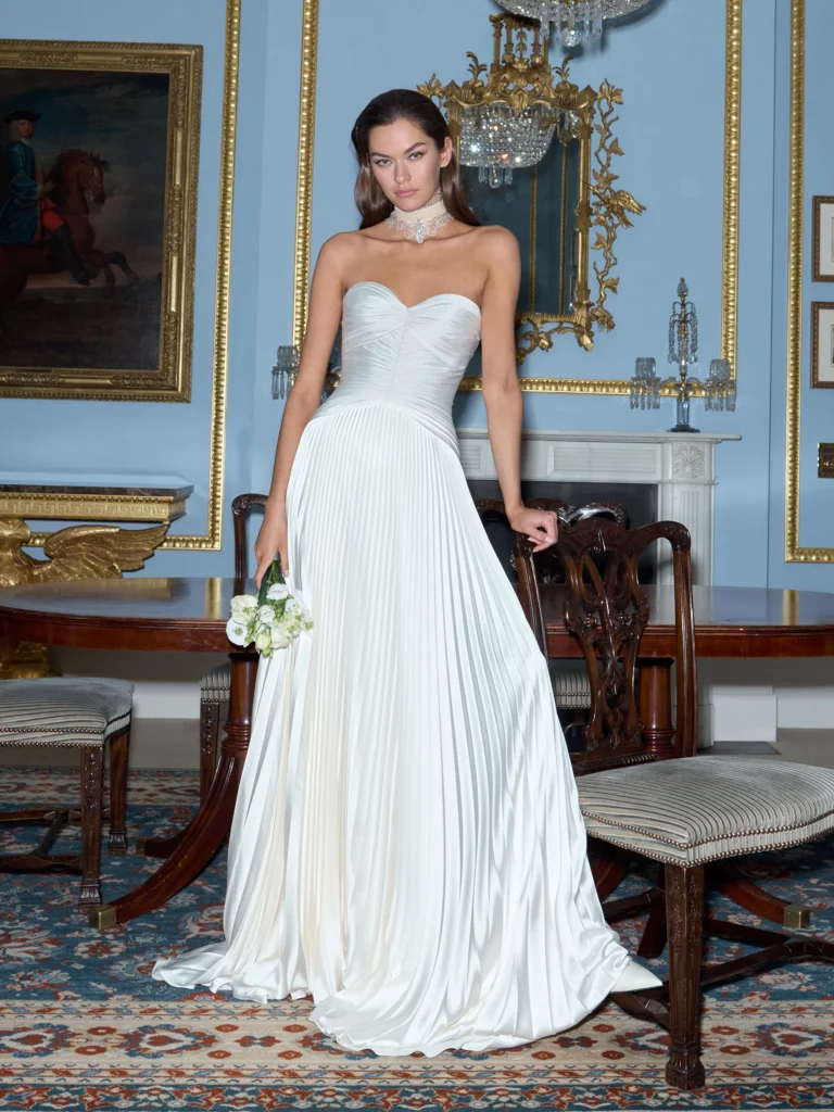 Model in a draped white gown with a sweetheart neckline, striking a pose indoors. A floral bouquet complements her elegant look against an ornate backdrop.