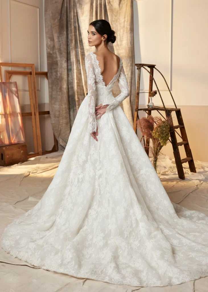 Elegant bride in a backless lace gown gazes over her shoulder, showcasing intricate details. Soft, neutral background complements the bridal aesthetics.