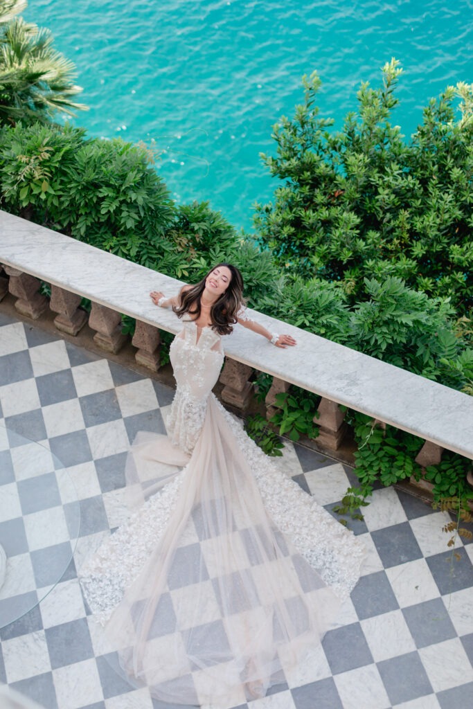 A woman in a stunning floral wedding gown poses joyfully on a balcony overlooking vibrant blue water, surrounded by lush greenery.