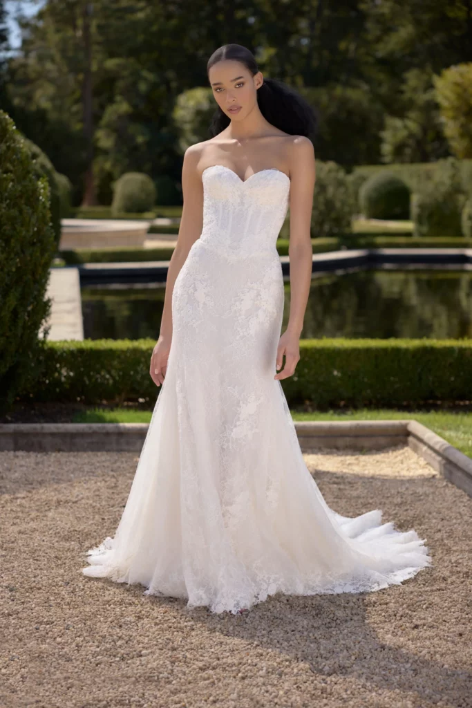 A model showcases a stunning strapless white wedding gown with intricate lace detailing, positioned in an elegant garden setting.