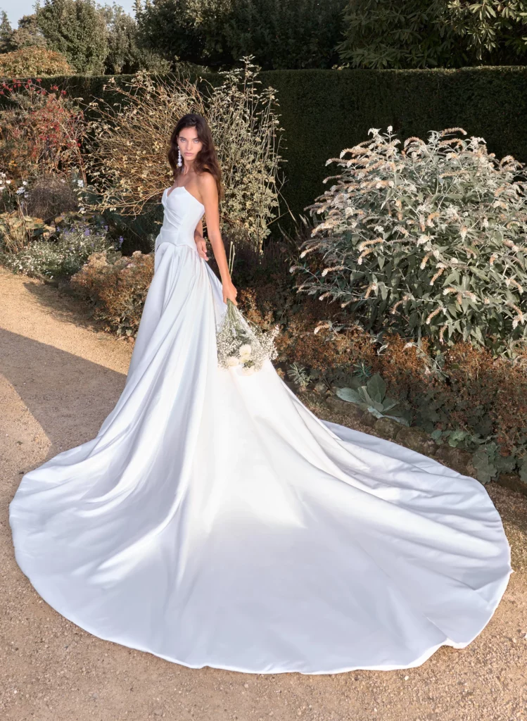 Bride in a flowing white gown stands in a garden, holding a bouquet of flowers, showcasing elegance and romance in a tranquil setting.