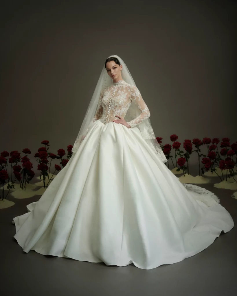 Bride in an elegant, intricately detailed wedding gown with a long veil, standing amidst a backdrop of red roses, exuding grace and romance.