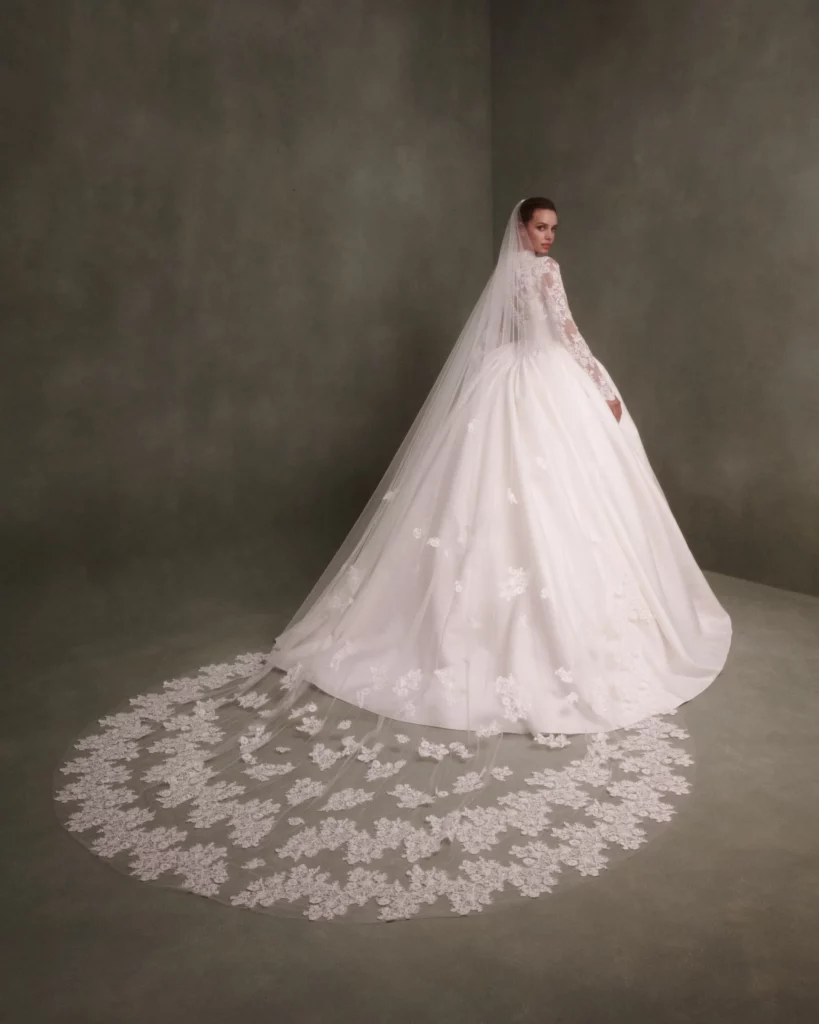 Bride in a white ball gown with intricate lace detailing and a long veil, showcasing a delicate floral train against a muted backdrop.