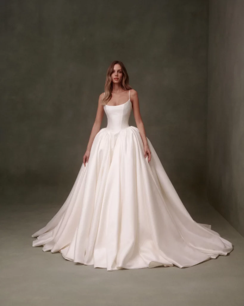 A model poses in a stunning, long white gown with a fitted bodice and voluminous skirt against a muted green backdrop, highlighting bridal fashion elegance.