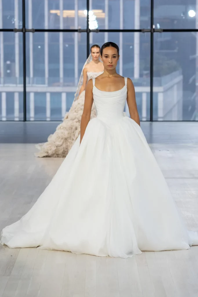 Model in a flowing white gown stands confidently in a modern bridal fashion show, with another model in a detailed gown walking behind her.