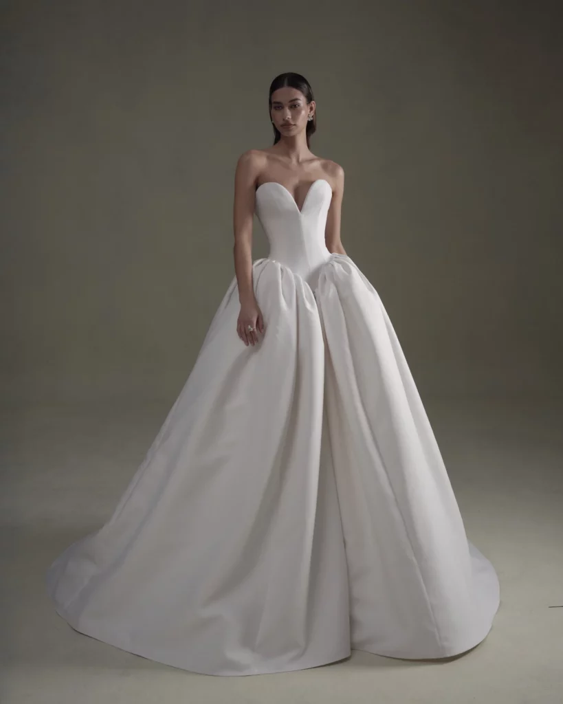 A model wears a stunning strapless white gown with a sweetheart neckline and voluminous skirt, showcasing modern bridal elegance.