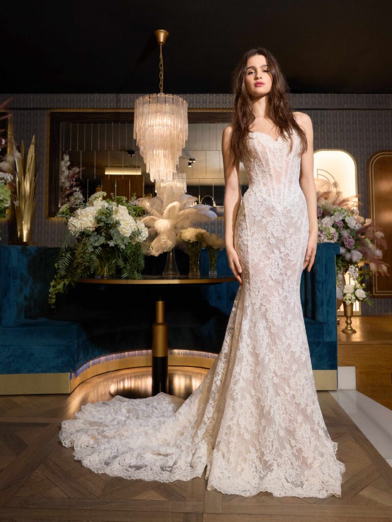 Model showcasing a delicate lace wedding gown with a long train, set in an elegant indoor space adorned with floral arrangements and a chandelier.